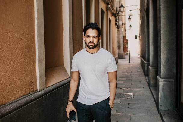 Content attractive hispanic man in white tshirt walking old town streets holding mobile phone hand.Bearded hipster male enjoying city travel on gotic district streets. - 写真・画像