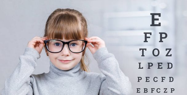Ein lächelndes Kind mit Brille steht vor dem verschwommenen Hintergrund des Arztzimmers, hinter dem ein Tisch mit Buchstaben steht. das Konzept der ophthalmologischen Diagnostik, Behandlung und Korrektur. - Foto, Bild