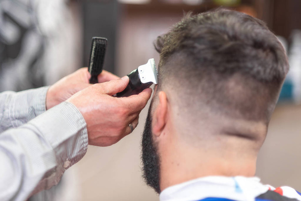 Hombre barbudo guapo, consiguiendo corte de pelo por peluquero, con trimmer eléctrico en la barbería
. - Foto, Imagen
