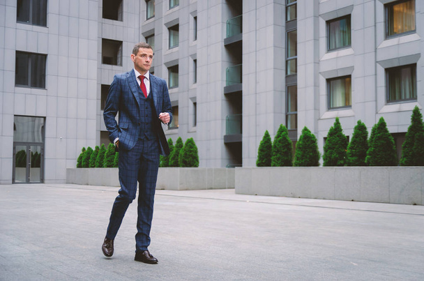 Stilvoller Geschäftsmann in schicker karierter Jacke, der in der Nähe eines betongrauen Geschäftszentrums spaziert. modernes Geschäftsleben - Foto, Bild