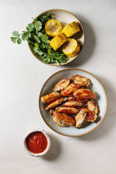 BBQ spicy chicken wings served in ceramic plate with red sauce, grilled sweet corn and parsley bundle in bowls over white marble background. Flat lay, space. - Foto, imagen