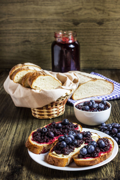 Sweet sandwiches with homemade blueberry jam and fresh blueberries. Slices of bread in a basket. Jam in a jar and a bowl in the background. White bread. Blueberries Wooden background. Rustik - Фото, зображення
