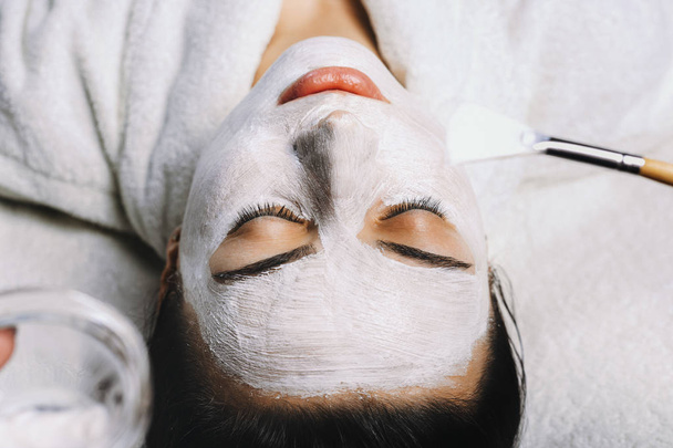 Hermosa mujer europea que tiene procedimiento de piel facial mediante la aplicación de una máscara de rejuvenecimiento blanco por el cosmetólogo
. - Foto, Imagen