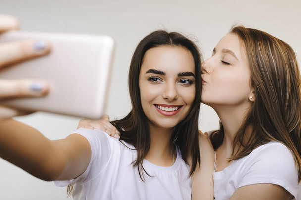 Two beautiful girlfriends doing selfie smiling while one is kissing another on the head in front of a white background. - Fotografie, Obrázek