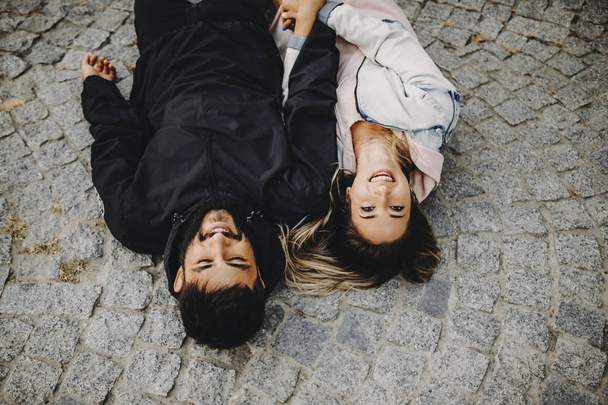 Lovely young caucasian couple having fun laughing and holding by hands while lying on the ground in the city. - Photo, Image