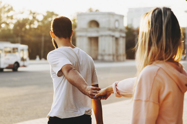 Задний вид на прекрасную пару, где мужчина держит ее подругу за руку и показывает ей, куда идти через город
. - Фото, изображение