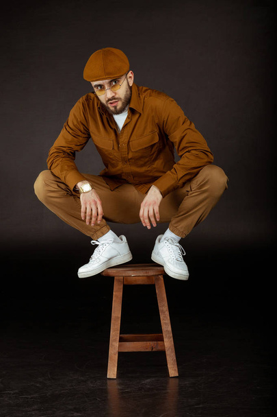 Stylish young man in beige clothes and yellowe sunglasses standing on chair - Photo, Image