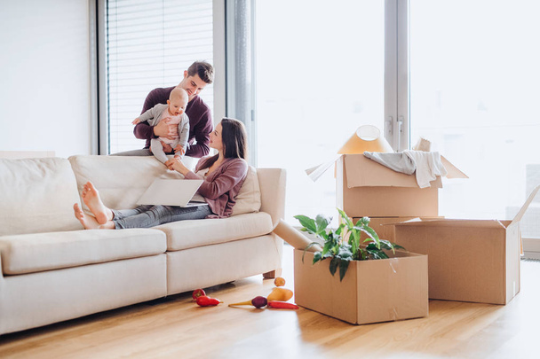 A portrait of young couple with a baby and cardboard boxes moving in a new home. - Foto, Imagen