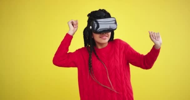 Very pretty african lady using a VR moving charismatic dacing in a studio with a yellow background, she enjoying the time with a virtual reality glasses - Imágenes, Vídeo