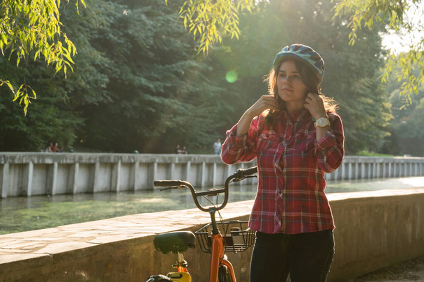 Θέμα οικολογική λειτουργία μεταφοράς ποδηλάτων. Όμορφη νεαρή kasazy γυναίκα, φορώντας ένα μπλε κράνος και μακριά μαλλιά θέτει στέκεται δίπλα σε ένα ποδήλατο ενοικίαση πορτοκαλί χρώμα με ένα καλάθι σε ένα πάρκο της πόλης. - Φωτογραφία, εικόνα