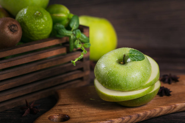 Concept of healthy food. Sliced green apple on cutting board and fresh green fruits in a box. Dark wooden background. Set of green fruits for healthy diet: apple, lime, kiwi, carambola and mint - Fotó, kép