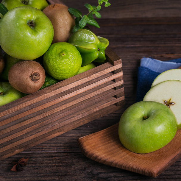 Concept of healthy food. Fresh green fruits in a box, green apple on cutting board. Dark wooden background. Set of green fruits for healthy diet and detox: apple, lime, kiwi, mango, carambola and mint - Foto, Imagem