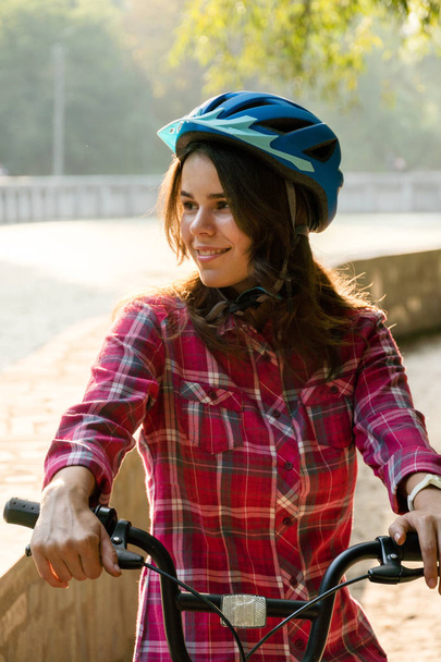 Subject ecological mode of transport bicycle. Beautiful young kasazy woman wearing a blue helmet and long hair poses standing next to an orange-colored rental bike with a basket in a city park. - Photo, Image