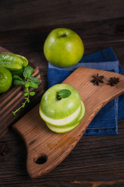 Concept of healthy food. Sliced green apple on cutting board and fresh green fruits in a box. Dark wooden background. Set of green fruits for healthy diet and detox: apple, lime, carambola and mint. - Photo, Image