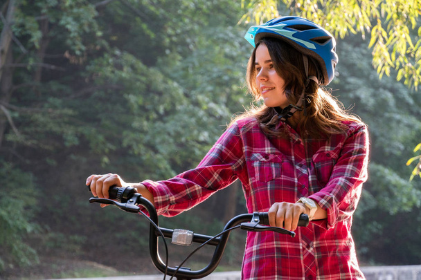 Onderwerp ecologische wijze van vervoer-fiets. Mooie jonge kasazy vrouw droeg een blauwe helm en lange haren vormt die naast een oranje-gekleurde huurfiets met een mandje in een stadspark. - Foto, afbeelding