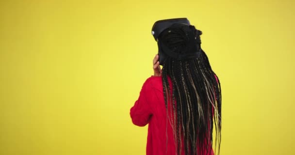 In the studio with a yellow background multi ethnic lady , with long hair dreadlocks enthusiastic exploring the game or traveling through the virtual reality glasses - 映像、動画