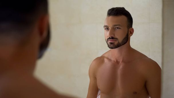 Bearded handsome man looking in mirror, perfect body, daily workout result - Фото, изображение