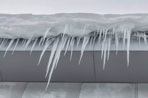 Eine Gruppe scharfer weißer Eiszapfen hängt vom grauen Dach eines Gebäudes herunter - Foto, Bild