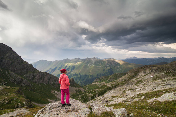 view from the back on girl in pink pants standing on a cliff  hands in pockets on a mountain background - Fotoğraf, Görsel