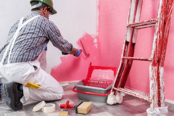 Peintre de maison caucasien travailleur en salopette de travail blanc, avec le rouleau, il peint le mur avec la peinture colorée de rose. Industrie de la construction. Sécurité au travail. Vue du bas
. - Photo, image