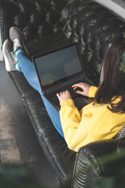 Photo recadrée verticale d'une dame méconnaissable travaillant sur un ordinateur portable à l'intérieur d'un café. Elle est allongée sur un canapé ou un canapé confortable et écrit une lettre à ses amis
 - Photo, image