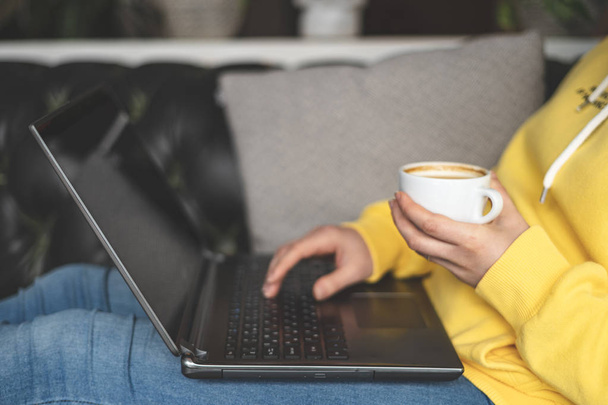 Profile side view cropped photo of unrecognizable person in trend trendy wear holding cup of coffee and writing text on laptop. She lying on comfort couch or sofa inside lovely quiet place - Фото, изображение