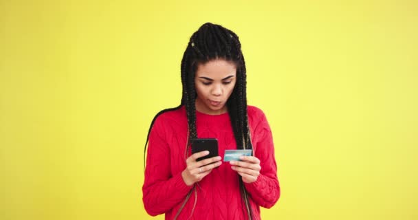In a studio with yellow background wall african lady in a red sweater in front of the camera using a smartphone and bank card to order something - Metraje, vídeo