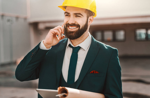 Portrait of bearded architect in formal wear talking on the phone and holding project and tablet while standing on construction site. - Photo, Image