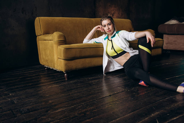 Full length portrait of a young fitness woman in sportswear posing near a yellow sofa sitting on the floor - 写真・画像