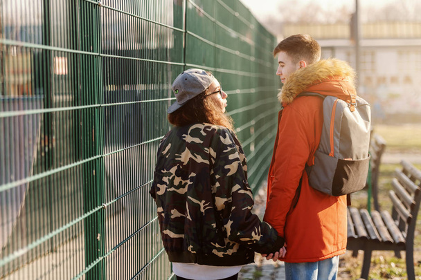 Cute teenage couple holding hands and walking next to fence. Backs turned. - Zdjęcie, obraz