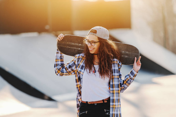 Όμορφη χαμογελώντας Μικτή φυλή εφηβικό αστικών κορίτσι με σγουρά μαλλιά που θέτουν με skateboard στο πάρκο skate. - Φωτογραφία, εικόνα