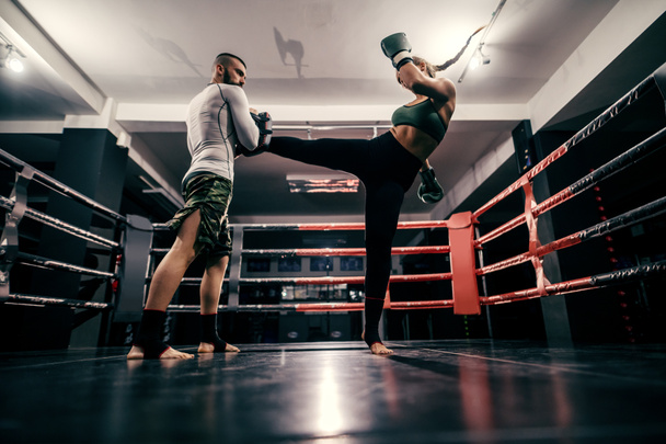 Powerful caucasian boxer girl in sportswear, with bandage on feet and boxing gloves on hands practicing with trainer in ring. - Photo, Image