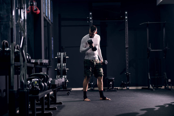 Мускулистый кавказский боксер в спортивной одежде и бинтах на ногах, поднимающий гантели в спортзале
. - Фото, изображение