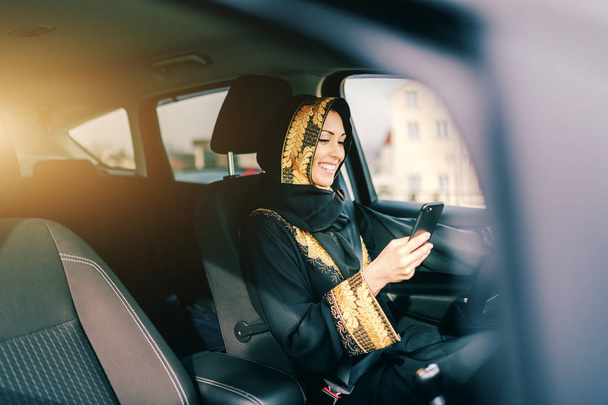 Портрет красивої усміхненої мусульманки з шарфом на голові за кермом автомобіля і за допомогою смартфона
. - Фото, зображення