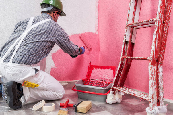 Καυκάσιος σπίτι ζωγράφος εργαζόμενος σε άσπρο εργασίας ολόσωμες φόρμες, με τον κύλινδρο που ζωγραφίζει στον τοίχο με τη χρωματισμένη ζωγραφική του ροζ. Κατασκευαστική βιομηχανία. Ασφάλεια εργασίας. Κάτω όψη. - Φωτογραφία, εικόνα
