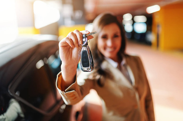 Close up de sorrir bela morena segurando chaves do carro no estacionamento. Foco seletivo na mão com teclas
. - Foto, Imagem