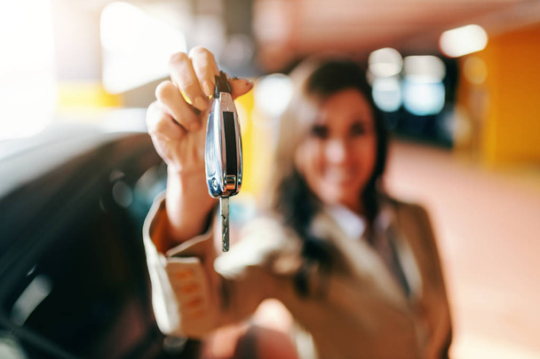 Close up of smiling beautiful brunette holding car keys at parking. Selective focus on hand with keys. - Foto, Imagen