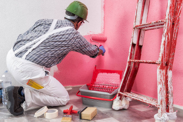Kaukasische huisschilder werknemer in witte overalls werken, met de roller schildert hij de muur met de gekleurde schilderij van roze. Bouwsector. De veiligheid van het werk. Onderaanzicht. - Foto, afbeelding