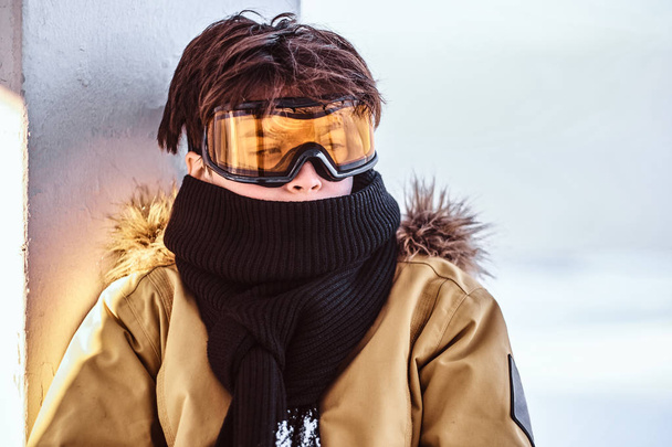 A teenage snowboarder dressed in snowsuit and protective goggles sitting on railing near the snowy beach - Foto, Imagem