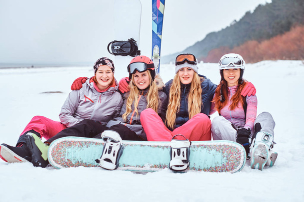 Друзья-женщины в спортивной зимней одежде с сноубордами и лыжами, обнимающимися вместе и смотрящими в камеру, сидя на снежном пляже
 - Фото, изображение
