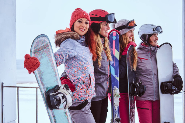 Портрет друзів-жінок у спортивному зимовому одязі, що тримає сноуборди та лижі, що стоять разом біля засніженого морського узбережжя
 - Фото, зображення
