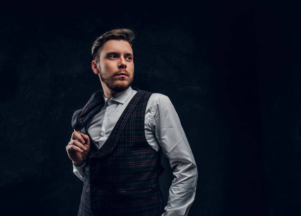 Elegantly dressed man holds jacket on the shoulder while posing against a dark textured wall - Foto, immagini