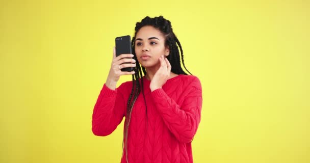 Beautiful african young woman using her smartphone to take a photo of her self in the studio with a yellow background wall in front of the camera she looks very carefully at her face and posing pretty - Video, Çekim