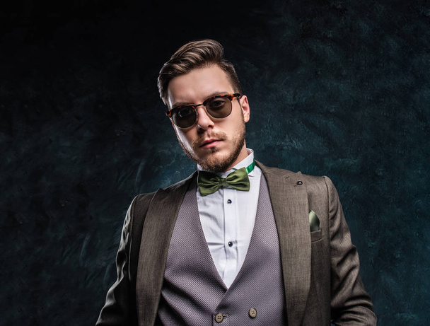 A stylish young man in sunglasses dressed in an elegant suit against a dark textured wall - Photo, image