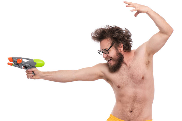 Crazy bearded Man with funny Haircut in eye Glasses, ready for fun at sunny beach. Happy and silly tourist, isolated on white background. Cheerful naked man holding Water Gun. - Фото, изображение