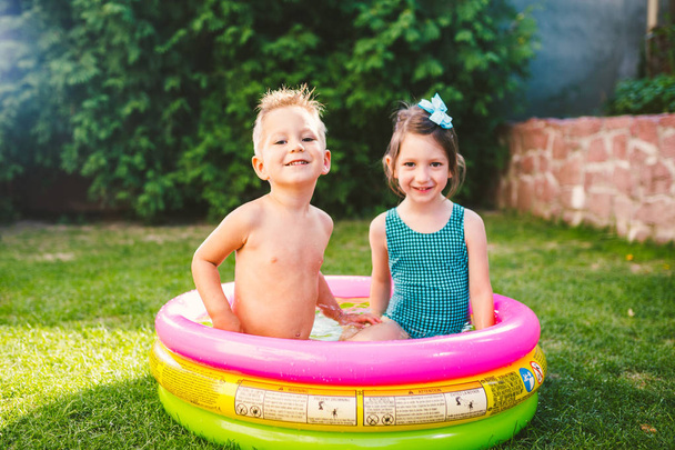 Thema sind die Sommerferien der Kinder. Zwei kaukasische Kinder, Bruder und Schwester, sitzen in Badeanzug und fröhlichem Lächeln in einem runden Pool mit Wasser im Hof des grünen Rasens.. - Foto, Bild