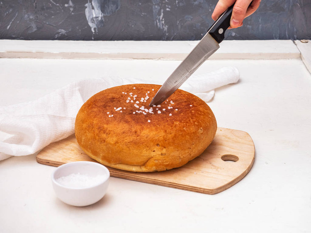 A knife in a human hand cuts freshly baked homemade bread. Bread lies on a wooden kitchen board, near the salt shaker with coarse salt. White wooden table. - 写真・画像