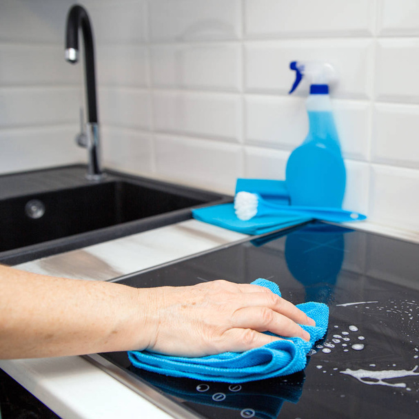 Die Hand einer Frau mit einem blauen Mikrofasertuch reibt in der Küche eine Glaskeramikplatte. Im Hintergrund sind Reinigungsprodukte. - Foto, Bild