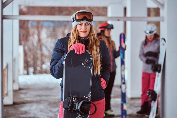 Norwegian woman wearing warm clothes and goggles posing with a snowboard in the winter ski resort - Fotó, kép