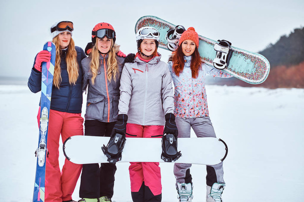 Women friends in sports winter clothes with snowboards and skis standing together in a hug and looking at the camera - Photo, Image
